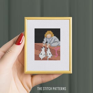 May include: A framed cross-stitch artwork featuring a person with two dogs. The artwork has a gold frame and is held by a hand with red nail polish. The text "THE STITCH PATTERNS" is at the bottom of the frame.