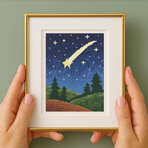 以下が含まれることがあります： 夜空のシーンを描いた、額装されたクロスステッチのアートワーク。 黄色の流れ星、白い星、緑の木々が濃い青色の背景に描かれています。 アートワークは、白いマットが付いた金色のフレームに額装されています。