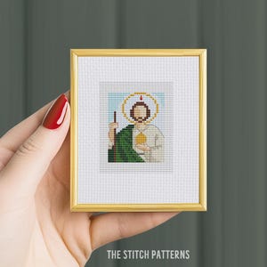May include: A small framed cross-stitch pattern of a saint with a halo, holding a staff and a heart. The saint is wearing a green robe and a white tunic. The background is a light blue sky.