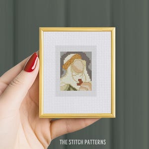 Op de afbeelding: Een ingelijst kruissteekpatroon van een persoon met een hoed die een rood voorwerp vasthoudt. Het kruissteek is op witte stof met een gouden lijst.