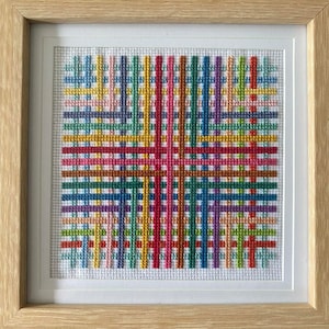Op de afbeelding: Een ingelijst kruissteekkunstwerk met een kleurrijk geometrisch patroon van elkaar kruisende lijnen in tinten rood, oranje, geel, groen, blauw, paars en roze.