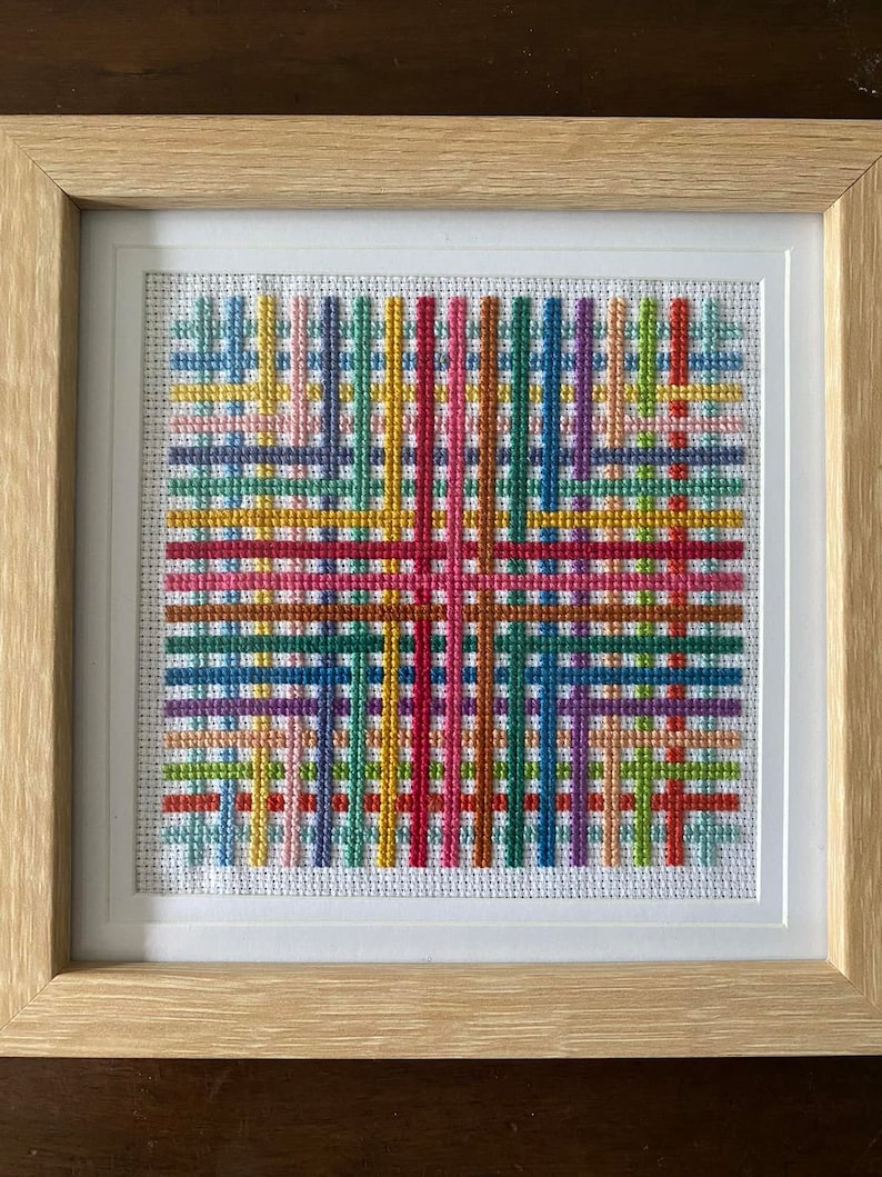 Op de afbeelding: Een ingelijst kruissteekkunstwerk met een kleurrijk geometrisch patroon van elkaar kruisende lijnen in tinten rood, oranje, geel, groen, blauw, paars en roze.
