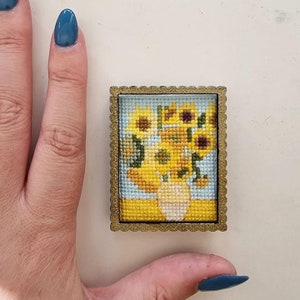 May include: A small framed cross-stitch of a vase of sunflowers. The sunflowers are yellow with brown centers and green leaves. The vase is white and the background is light blue.