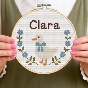 Könnte beinhalten: Ein Kreuzstich-Stickrahmen mit dem Namen "Clara" in Braun über einer Ente mit einer blauen Schleife. Die Ente ist von einem floralen Kranz aus blauen Blumen und grünen Blättern umgeben. Der Rahmen wird von Händen mit rosa Nagellack gehalten.