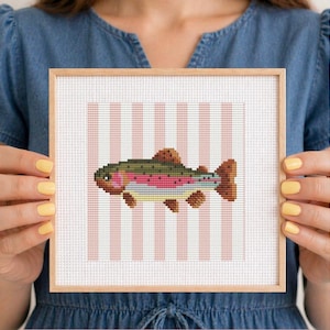 May include: A square cross-stitch embroidery featuring a rainbow trout with green, pink, and yellow scales centered on a white fabric background with vertical light pink stripes, all enclosed within a simple light-colored wooden frame.