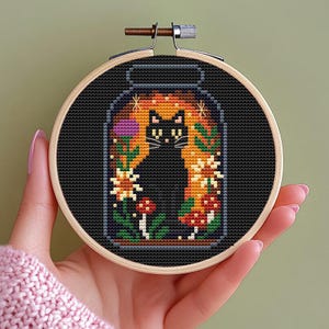 May include: A cross-stitch embroidery hoop featuring a black cat inside a jar-shaped design. The cat is surrounded by colorful flowers, mushrooms, and foliage, set against a black background. The hoop is held in a hand.
