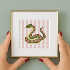 May include: A framed cross-stitch art piece featuring a green snake with pink polka dots on a pink and white striped background. The artwork is displayed in a gold-colored frame.
