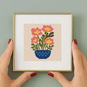 May include: A framed cross-stitch artwork featuring a floral design. The artwork showcases four pink and yellow flowers in a blue pot against a beige background. The frame is gold-colored.