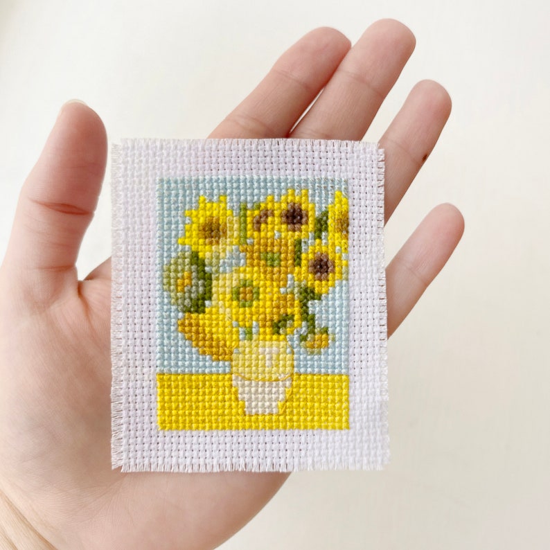 May include: A small, square cross-stitch of sunflowers in a white vase on a yellow table, set against a light blue background. The artwork is on white fabric with a frayed edge. The piece is held in a hand.