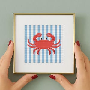 May include: A framed cross-stitch art piece featuring a red crab on a blue and white striped background. The crab has black eyes and is centered in the frame. The frame is gold-colored.