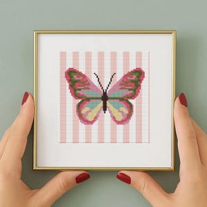 Könnte beinhalten: Gerahmtes Kreuzstich-Kunstwerk mit einem bunten Schmetterling mit rosa, grünen und gelben Flügeln. Der Hintergrund ist mit rosa und weißen Streifen versehen. Der Rahmen ist goldfarben.