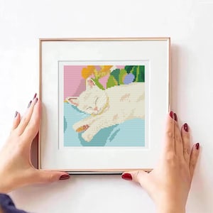Könnte beinhalten: Gerahmtes Kreuzstich-Kunstwerk mit einer schlafenden weißen Katze auf einer blauen Oberfläche. Der Hintergrund zeigt gelbe und grüne Blumenelemente vor einem rosa Hintergrund. Der Rahmen ist goldfarben.