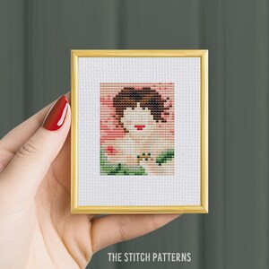 May include: A cross-stitch pattern of a woman with brown hair and red lips, wearing a green dress and surrounded by pink flowers.