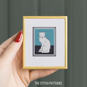 Puede incluir: Un patrón de punto de cruz de un gato blanco en un marco dorado. El gato está sentado con los ojos cerrados y está delineado en negro. El fondo es un bloque de color azul y negro.