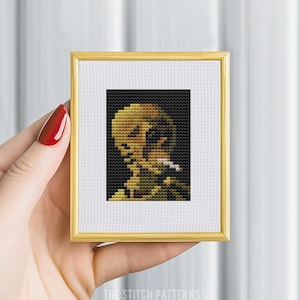 May include: A cross-stitch pattern of a portrait of a person with a cigarette in their hand. The person is depicted in a yellow and brown color scheme against a black background.