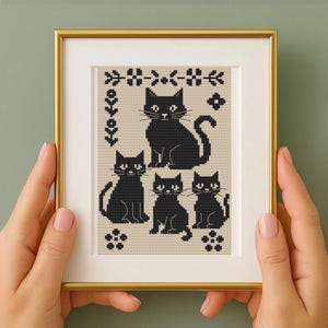 Könnte beinhalten: Ein gerahmtes Kreuzstich-Kunstwerk mit fünf schwarzen Katzen in verschiedenen Posen vor einem beigefarbenen Hintergrund. Die Katzen sind von floralen und geometrischen Mustern umgeben, alles in einem goldfarbenen Rahmen. Das Kunstwerk wird von einer Person gehalten.