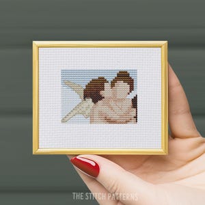 Puede incluir: Una bordadura de punto de cruz enmarcada que representa a dos personas abrazándose. La bordadura está hecha en tonos de marrón, rosa y blanco sobre un fondo azul claro.