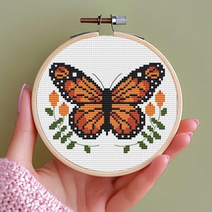 May include: A cross-stitch embroidery hoop featuring a monarch butterfly design. The butterfly has orange and black wings, with a white background. The design includes orange flowers and green leaves. The hoop is made of wood.
