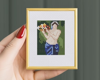 May include: A small cross-stitch embroidery in a gold frame featuring a figure holding a basket of fruit against a green background. The figure wears a blue patterned lower garment and has a white head covering. The text THE STITCH PATTERNS is visible.