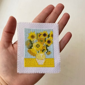 May include: A cross-stitch embroidery of a vase of sunflowers. The sunflowers are yellow with brown centers and green leaves. The vase is white and the background is blue and yellow.