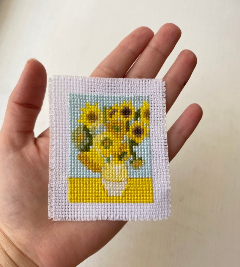 May include: A cross-stitch embroidery of a vase of sunflowers. The sunflowers are yellow with brown centers and green leaves. The vase is white and the background is blue and yellow.