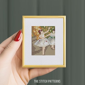 May include: A framed cross-stitch pattern of a ballerina in a white tutu with a red heart on her chest. The ballerina is surrounded by a colorful background.