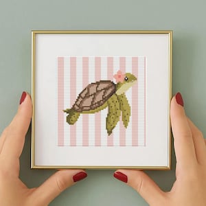 May include: A framed cross-stitch art piece featuring a green sea turtle with a brown shell and a pink flower on its head. The background has pink and white vertical stripes. The frame is gold.