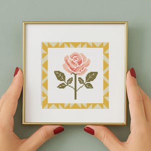 May include: A framed cross-stitch of a pink rose with green leaves and stem. The artwork is set against a white background with a yellow geometric border. The frame is gold-colored.