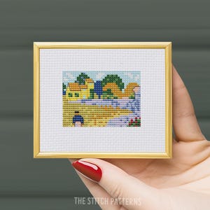 May include: A cross-stitch pattern of a yellow house with a blue roof, green trees, and a person in a blue dress. The pattern is framed in a gold frame.