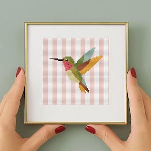 Puede incluir: Una pieza de arte de punto de cruz enmarcada con un colibrí colorido. El pájaro tiene la garganta roja, la cabeza verde y las alas azules, amarillas y marrones. El fondo tiene rayas verticales rosas y blancas. El marco es dorado.