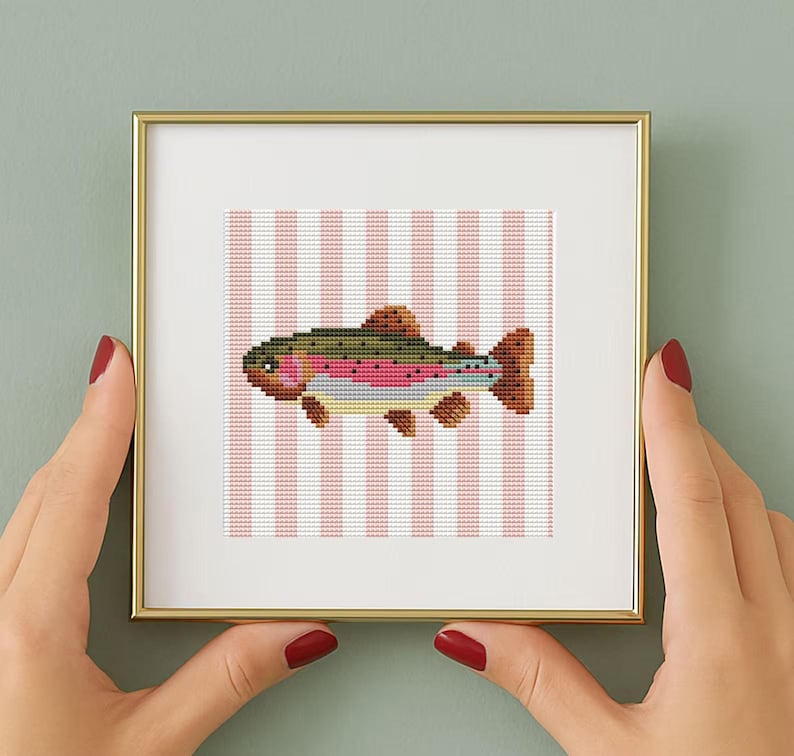 May include: A square framed cross-stitch embroidery featuring a rainbow trout fish with green, pink, and brown thread details. The fish is centered against a background of vertical white and light pink stripes, all encased in a thin gold frame.