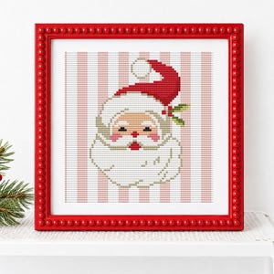 May include: A square red beaded frame containing a cross-stitch pattern of a Santa Claus face with a red hat, white beard, and pink cheeks, set against a background of vertical light pink and white stripes with a small green holly sprig.