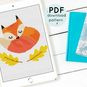 May include: A cross-stitch pattern featuring a sleeping fox with orange and white fur, curled up with its tail around its body. The pattern is surrounded by two yellow leaves.