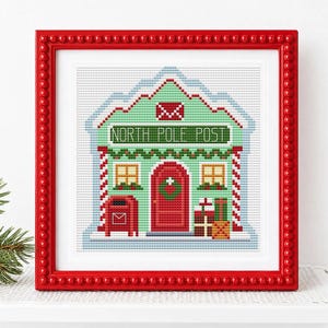 May include: A framed cross-stitch embroidery depicting a light green building labeled NORTH POLE POST. The design features a red door, a red mailbox, two windows, and stacked gift boxes, all enclosed in a bright red beaded frame.