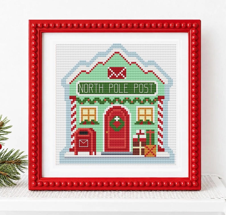 May include: A framed cross-stitch embroidery depicting a light green building labeled NORTH POLE POST. The design features a red door, a red mailbox, two windows, and stacked gift boxes, all enclosed in a bright red beaded frame.