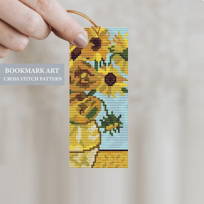 May include: A cross stitch bookmark pattern featuring a colorful sunflower design. The bookmark is made of fabric and has a brown string attached to the top.