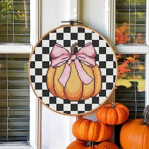 May include: A cross-stitch embroidery hoop with a black and white checkered background and a pumpkin with a pink bow in the center.