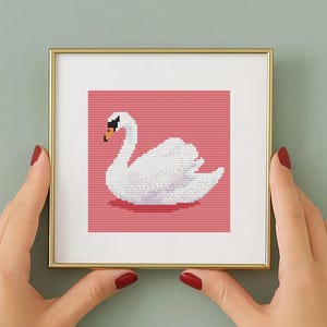 May include: A framed cross-stitch artwork featuring a white swan on a pink background. The swan has a black beak and is set within a gold-colored frame. The artwork is held by two hands with red nail polish.