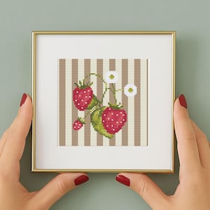 May include: A framed cross-stitch artwork depicting a strawberry plant with red berries, green leaves, and white flowers. The background has vertical brown and white stripes. The frame is gold-coloured.