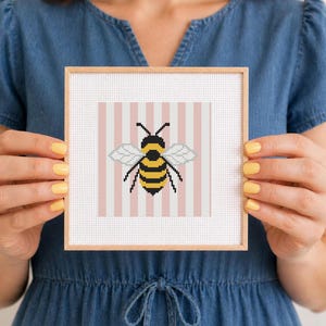 May include: A framed cross-stitch featuring a bee design. The bee is yellow and black, with gray wings, set against a pink and white striped background. The frame is a light wood color.