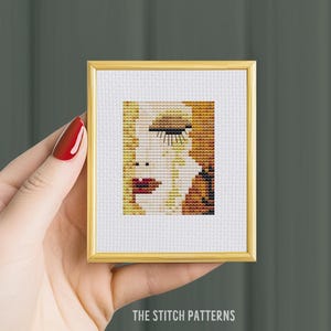 May include: A framed cross-stitch pattern of a woman's face with a closed eye, long eyelashes, and red lips. The background is a mix of yellow and orange.