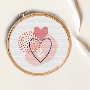 Puede incluir: Un patrón de punto de cruz con dos corazones, uno rojo y otro azul, sobre un fondo rosa y naranja. Los corazones están rodeados por un patrón de pequeños cuadrados rojos.