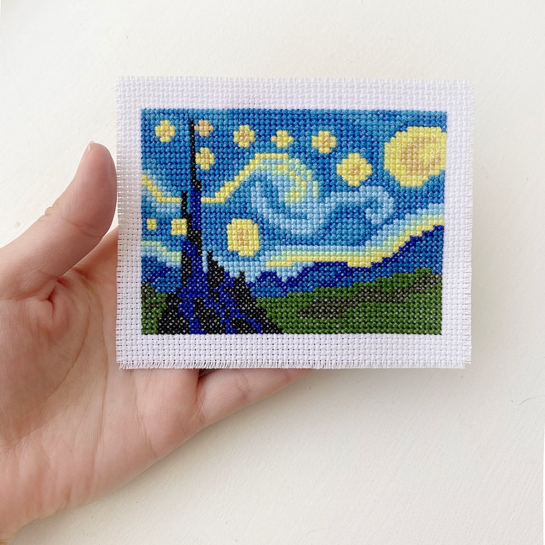 May include: A small, rectangular cross-stitch piece depicting Van Gogh's "Starry Night." The artwork features a dark blue sky with yellow stars and a crescent moon, above a dark silhouette of a town and green hills. The piece is mounted on white fabric.