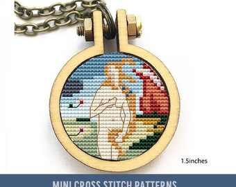 May include: A small, round wooden hoop with a cross-stitch design of a nude figure standing in front of a colorful landscape. The hoop is 1.5 inches in diameter and is attached to a chain necklace. "MINI CROSS STITCH PATTERNS" is printed below the hoop.