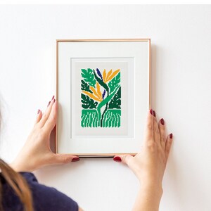 May include: A framed cross-stitch artwork featuring a stylized bird of paradise flower with green, yellow, and blue colors.
