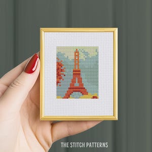 May include: A framed cross-stitch pattern of the Eiffel Tower in Paris, France. The tower is red and the background is a blue sky with white clouds.