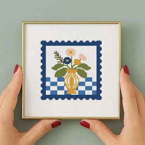 May include: A framed cross-stitch artwork featuring a yellow vase with flowers. The artwork has a blue and white checkered background and a blue border. The frame is gold.
