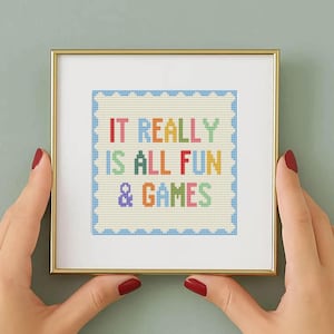Könnte beinhalten: Ein gerahmtes Kreuzstich-Kunstwerk mit dem Text "IT REALLY IS ALL FUN & GAMES" in bunten Buchstaben. Das Kunstwerk befindet sich in einem goldenen Rahmen, der von Händen mit rotem Nagellack gehalten wird.