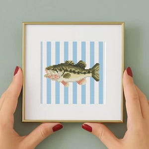 May include: A square framed cross-stitch artwork featuring a green and tan fish centered against a background of vertical light blue and white stripes. The piece is enclosed in a thin, metallic gold frame with a white mat border.