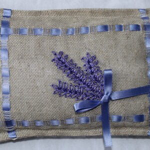 May include: A small, rectangular burlap pillow with a lavender flower design embroidered on the front. The pillow is trimmed with blue ribbon and has a bow on the front.